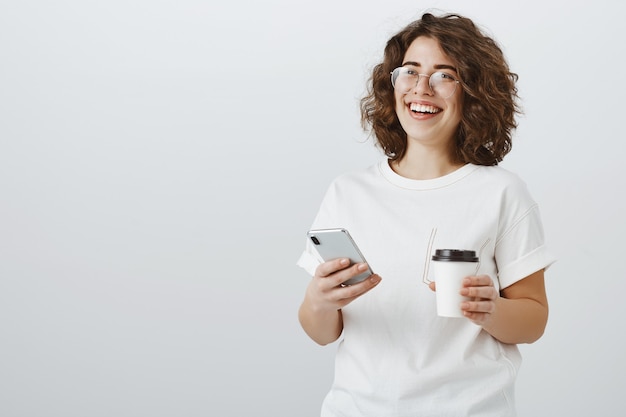 Женщина в очках, улыбаясь в текстовое сообщение мобильного телефона, пьет кофе во время перерыва в офисе