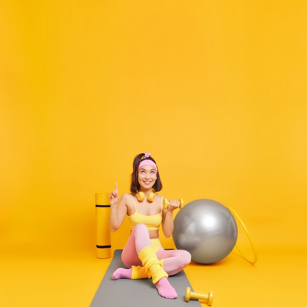 woman goes in for sport in fitness studio indicates upwards raises dumbbell demonstrates copy space 