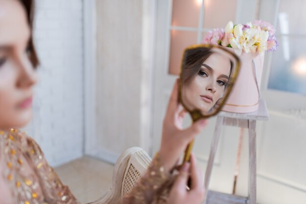 Woman in golden evening gawn and crown poses in luxury white room and looks in the mirror