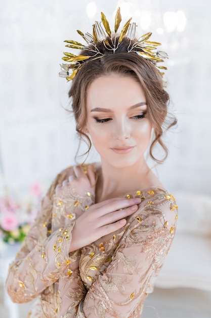 Woman in golden evening gawn and crown poses in luxury white room