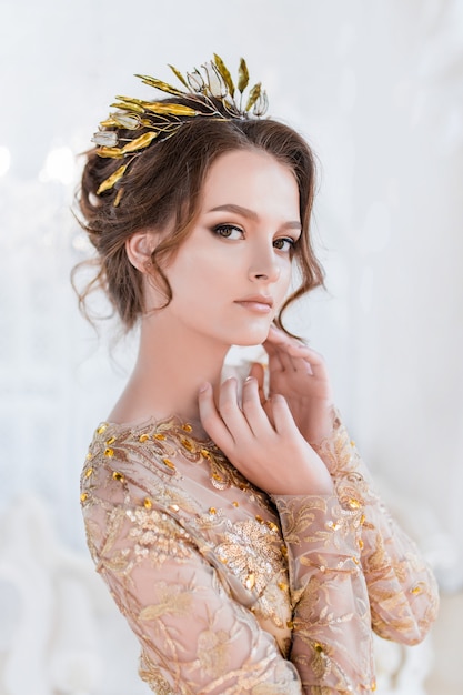 Woman in golden evening gawn and crown poses in luxury white room