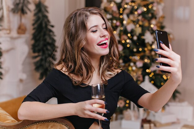 お祭り気分の女性は、電話でビデオ通話をして友達と話し、グラスワインを持っています。クリスマスの雰囲気の中で楽しいヨーロッパのモデルの肖像画