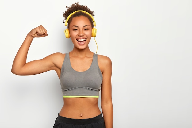 Woman in good mood, raises arm with muscles, has strong body, dressed in gym outfit, listens audio via modern headphones, poses indoor