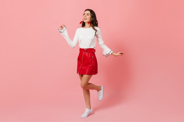 Woman in great mood runs against pink background and looks upwards admiringly. Lady in stylish leather skirt smiling.