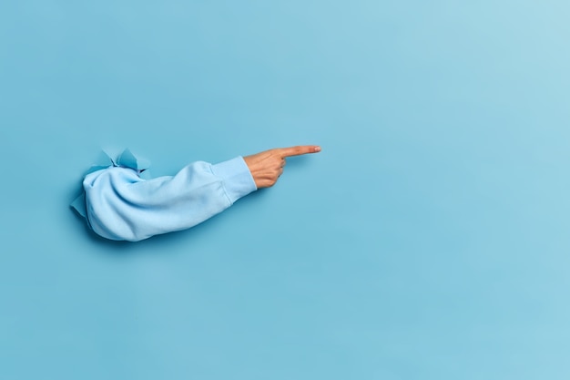 Woman hand in blue sweater breaking through paper wall and pointing to copy space