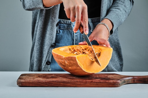 A woman hand cutting a slice of pumpkon with knife on wooden board .