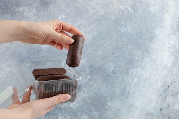 Woman hand holding a plastic box with chocolate cream rolls isolated on marble background.