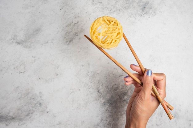 Woman hand holding raw tagliatelle pasta with wooden chopsticks on white background. 