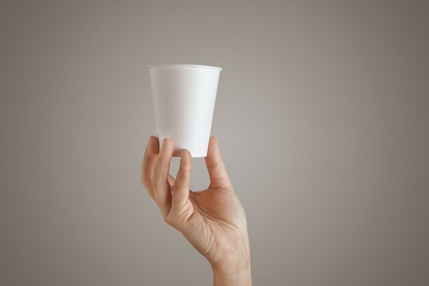 Woman hand holds blank empty take away paper glass from bottom, presentation in center, side view, Isolated, unrecognizable