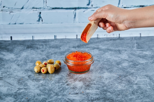 Woman hand taking a spoon of red caviar with olives on marble table.