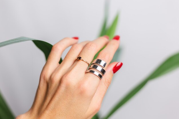 Woman hand with red manicure and two rings on fingers, on beautiful green palm leaf tropical. Gray wall behind.