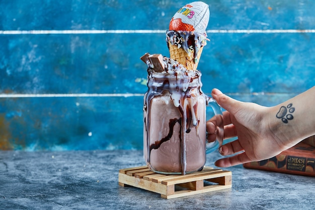 A woman hand with tattoo holding a mug of chocolate smoothie with kinder surprise, sprinkles in the blue surface