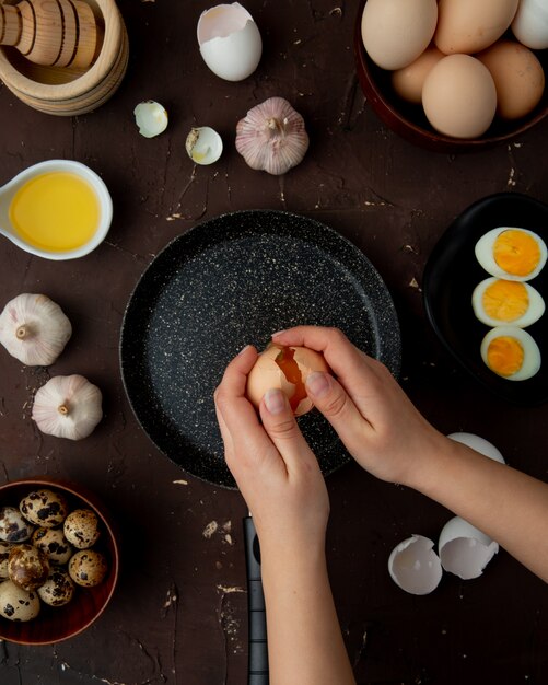  woman hands cracking open egg into frying pan and foods as butter garlic on maroon table