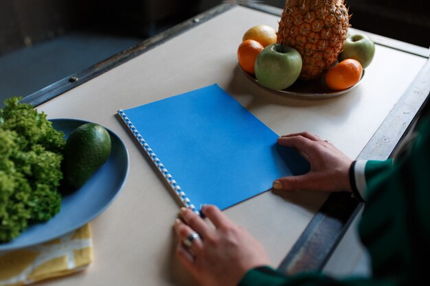 Woman hands holding notebook, on kitchen table with fruits and salad avocado. 