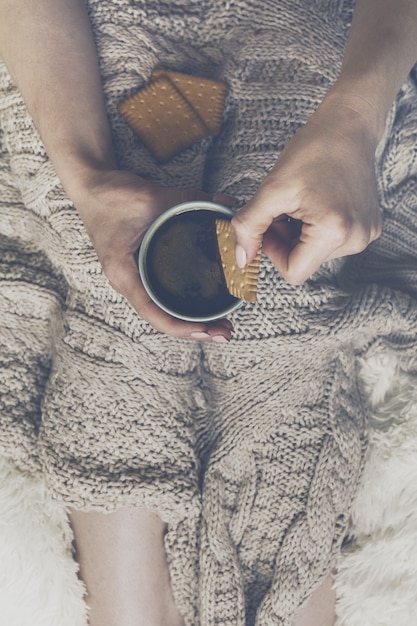 Free photo woman hands holding tasty warm coffee espresso in ceramic cup with cookie sitting on bed with plaid. home concept. top view.