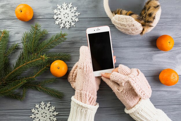Woman hands in mittens with smartphone near fir branches and paper snowflakes