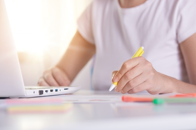 Woman hands with yellow pen writing something on peper, working online, female working on laptop