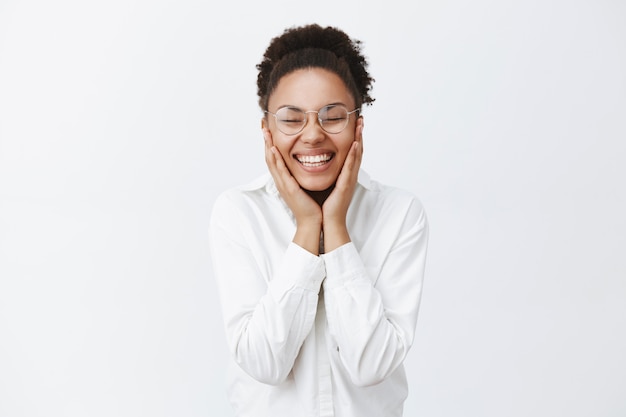 Woman happy to get rid of acne, touching beautiful and natural skin, holding palms on cheeks and smiling carefree from delight and pleasure, standing in glasses and white collar shirt over grey wall
