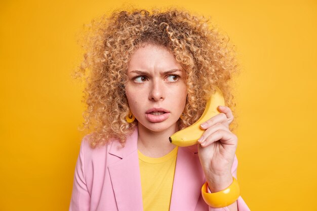 женщина сердится, строгое выражение лица играет роль босса, держит банан возле уха, притворяется, что телефонный звонок отвлечен, одетая в стильную куртку
