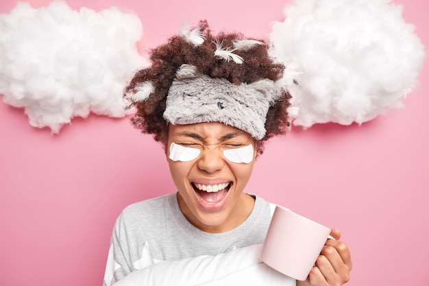 woman has curly Afro hair with stuck feathers screams loudly enjoys drinking morning coffee dressed in nightwear undergoes beauty procedures isolated on pink