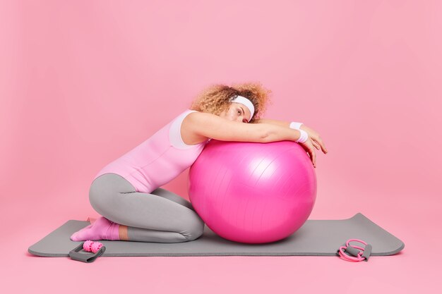 woman has exhausted look leans at fitness ball dressed in bodysuit uses sport equipment for workout 