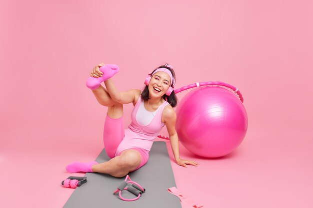 여성은 피트니스 매트를 올리고 다리를 쭉 뻗고 집에서 운동을 하며 헤드폰을 통해 음악을 듣습니다. 분홍색으로 격리된 활동복을 입은 스포츠 장비를 사용합니다