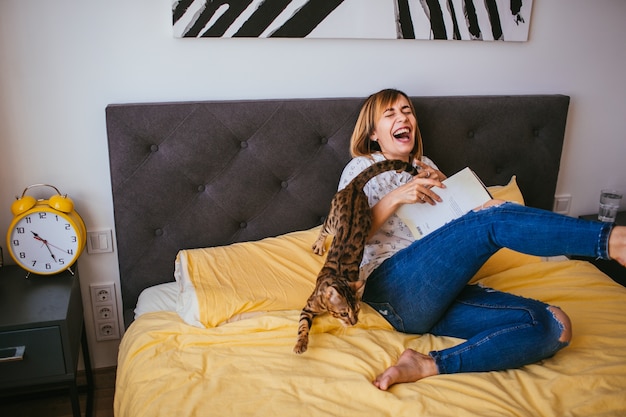 Woman has fun with Bengal cat on yellow bed 