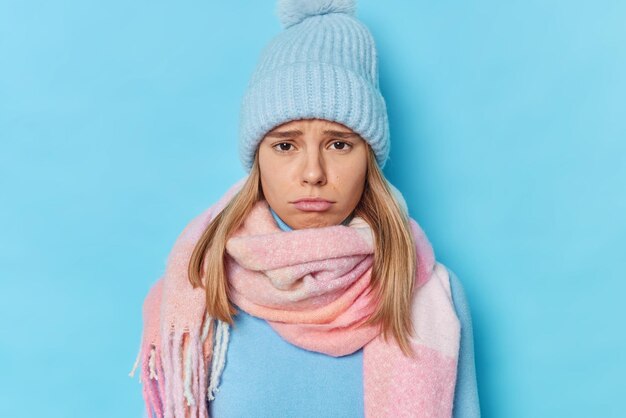 woman has gloomy expression as someone hurt her feelings complains about something wears knitted hat and scarf around neck isolated on blue feels guilty