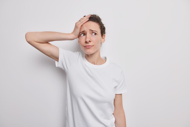 woman has headache worries because of life problems looks bothered keepss hand on head wears casual clothes poses on white. Oh no how can it be