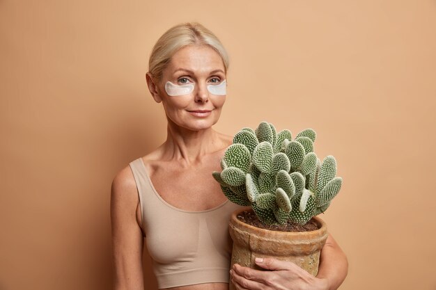 woman has pefect facial skin aplies beauty pads under eyes to remove wrinkles holds pot of cactus isolated on beige