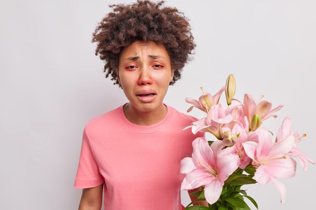woman has red swollen eyes reacts to allergen holds bouquet of lilies wears t shirt poses indoor