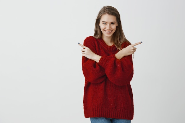 Woman has surprise, asking to pick direction. Indoor shot of good-looking european female in stylish loose red sweater, pointing in both sides with index fingers and smiling broadly over gray wall