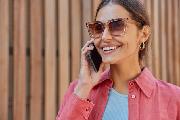  woman has telephone conversation wears trendy sunglasses and pink shirt feels happy smiles broadly poses on hence communicates via smartphone application