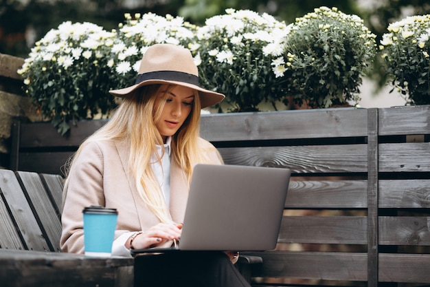 Woman in hat drinking coffee and working on laptop outside