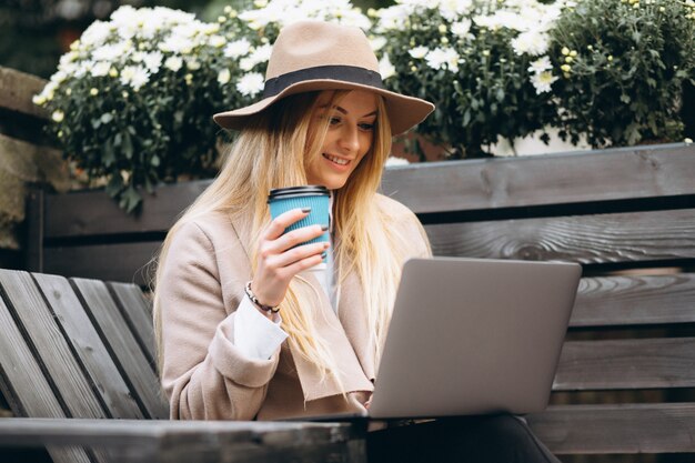 Woman in hat drinking coffee and working on laptop outside