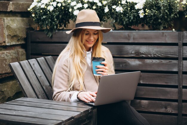 Woman in hat drinking coffee and working on laptop outside