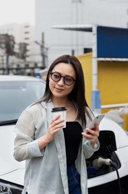 電気自動車の充電とスマートフォンの使用中にコーヒーブレイクをしている女性