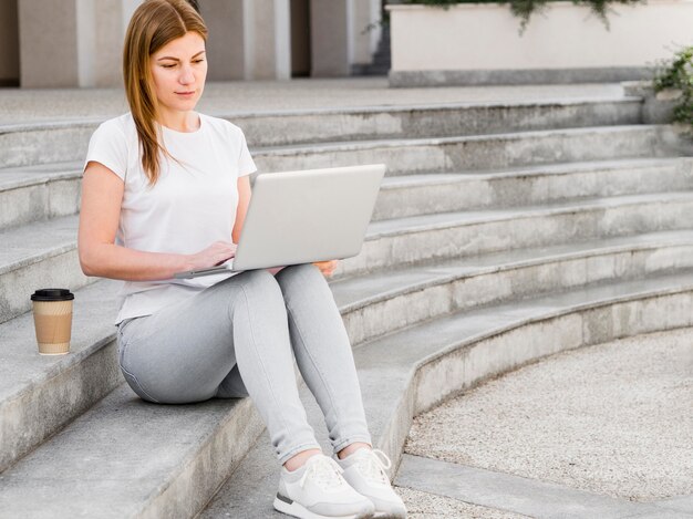 Woman having coffee on steps and working on laptop