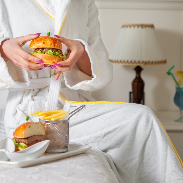 Woman having a meal, eating hamburger in bathrobe .