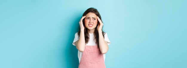Женщина с болезненной мигренью касается головы и хмурится, глядя в верхний левый угол, страдает головной болью, стоя на синем фоне