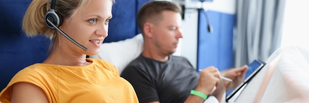 Woman in headphones lying in bed and working at laptop next to man with tablet remote work