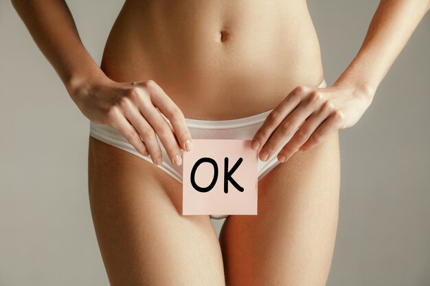 Woman health. Female model holding card near stomach. Young adult girl with paper with OK sign or symbol isolated on gray studio background. Cut out part of body. Medical problem and solution.