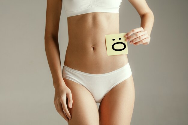 Woman health. Female model holding card with Emoji near stomach. Young adult girl with paper for sign or symbol isolated on gray studio background. Cut out part of body. Medical problem and solution.