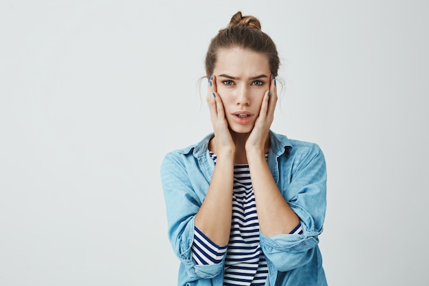 Woman heard horrible news and worries. Studio shot of young good-looking female student with troubled expression holding hands on face and staring , being shocked and nervous over gray wall