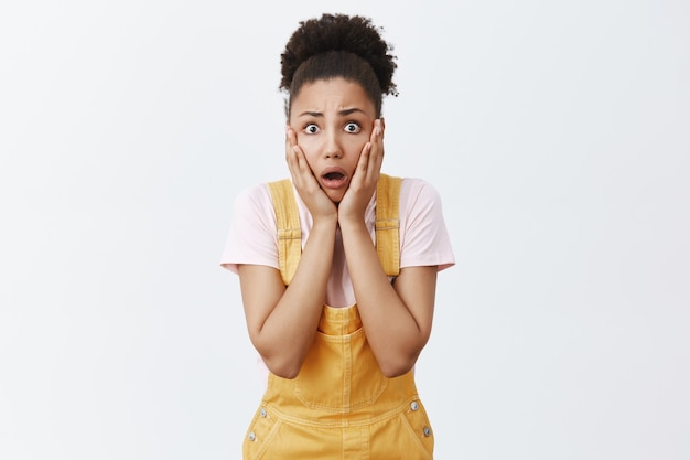 Woman hearing terrible disaster happened to friend. Portrait of shook intense worried female with dark skin in yellow overalls, gasping, losing speech and staring shocked, being astonished