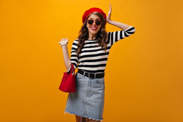 Woman in heart shaped sunglasses holds red handbag. Smiling cool girl with wavy hair in denim skirt and striped sweater posing.