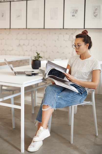 Woman in her twenties pursuing a career in fashion design looking at a magazine sitting in her bright studio with laptop computer.