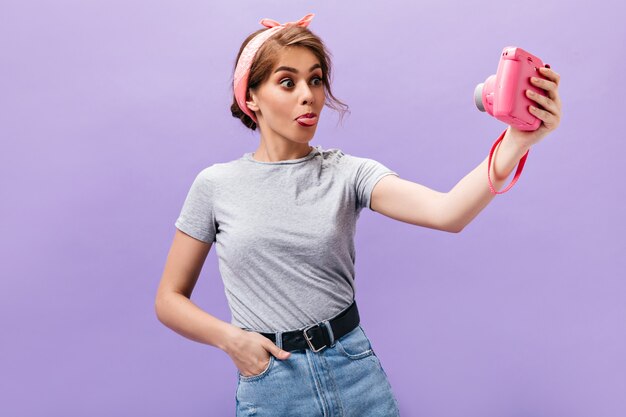 Woman in high spirit shows tongue and takes selfie. Funny wonderful lady in pink bandana and fashionable outfit posing.n purple background.