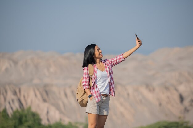 woman hiking in mountains standing on rocky summit ridge with backpack and pole looking out over landscape, happy female making self portrait in mountains