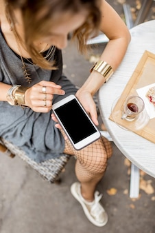 テーブル​の​上に​ケーキ​と​コーヒー​と​一緒​に​カフェ​で​屋外​に​座っている​空​の​画面​で​スマート​フォン​を​持っている​女性